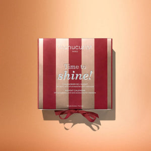 A red and gold Manucurist Advent Calendar Time to Shine! box with a ribbon, displayed on a peach background, featuring nail polishes, manicure accessories, and luxurious nail and hand care products like the innovative Active™ Shine Icy.